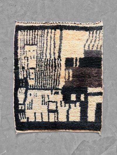 Alfombra marroquí Boujad de lana en negro y crema, con detalles en marrón chocolate, de diseño abstracto con rayas y bloques geométricos y cenefa negra. Vista general desde arriba.