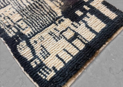 Alfombra marroquí Boujad en negro y crema. Vista general en diagonal sobre suelo gris, con el dibujo geométrico, la cenefa negra y el remate tejido.