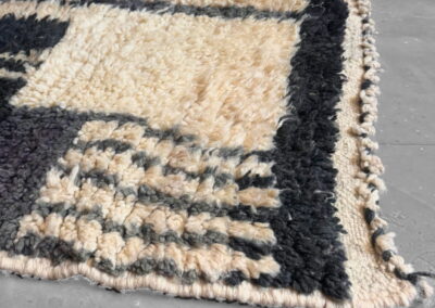 Detalle de alfombra marroquí Boujad en negro y crema. Esquina en perspectiva con la cenefa negra, el borde tejido en crema y los flecos trenzados.