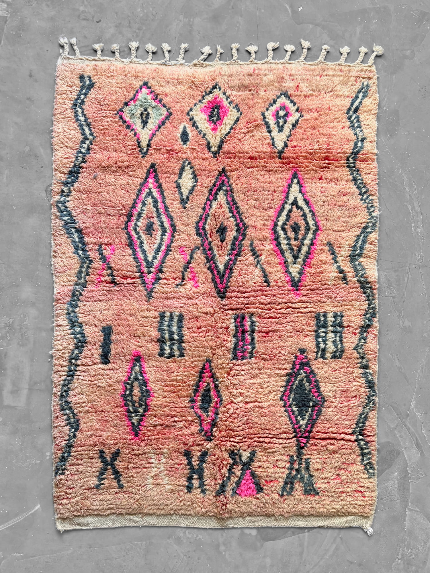 Alfombra marroquí Boujad New Works de lana en tonos rosa salmón, con rombos en gris antracita, fucsia y crudo, flecos anudados y remate de kilim crudo. Vista general.