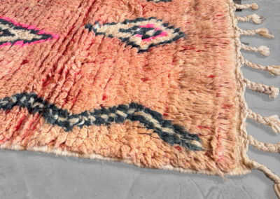 Alfombra marroquí Boujad New Works rosa salmón: esquina en perspectiva con rombos fucsia y antracita, zigzag lateral y flecos trenzados anudados. Detalle.