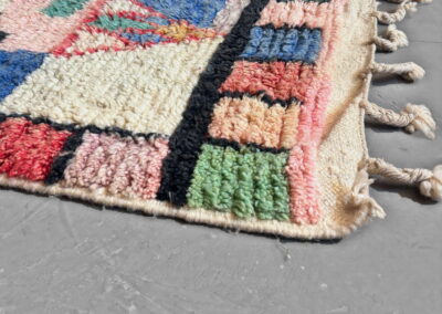 Detalle en perspectiva de una esquina de la alfombra Boujad nueva: borde de cuadros rosa, verde, azul y coral entre líneas negras, campo crema y rosa, y flecos trenzados de lana crema.