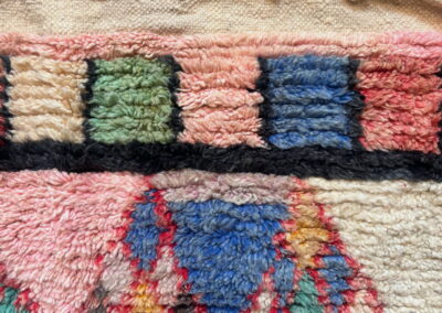 Detalle de un extremo de la alfombra Boujad nueva: flecos trenzados y franja tejida en crema, cenefa de cuadros verde, azul y coral, y triángulos azules, rosas y mostaza perfilados en rojo.