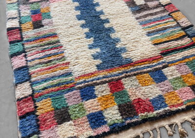 Vista general en perspectiva de la alfombra Boujad nueva: campo crema con motivo central azul, rayas multicolores a los lados, damero en rojo, mostaza, verde, azul y marrón, y flecos trenzados en los dos extremos.