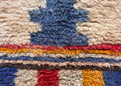 Detalle de cerca del pelo de lana de la alfombra Boujad nueva: motivo escalonado azul sobre fondo crema, franja de rayas roja y mostaza, y cuadros en rojo, verde, azul y mostaza.