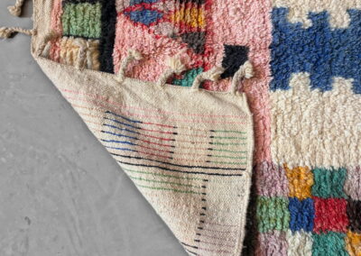 Detalle de una esquina doblada de la alfombra Boujad nueva que muestra el reverso tejido en crema con rayas finas de colores, junto al anverso en rosa, azul, verde y crema con flecos trenzados.