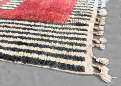 Detalle en perspectiva de alfombra Boujad con rayas gris antracita y crema, bloque rojo y flecos con borlas
